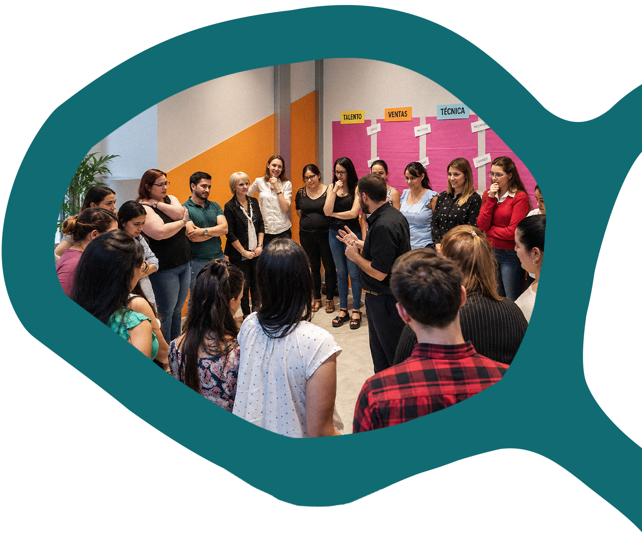 Grupo de participantes de pie en ronda escucha a un facilitador en una sala con carteles en la pared.